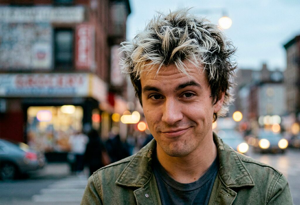 Young man with spiky frosted tips smirking on busy city street at dusk.