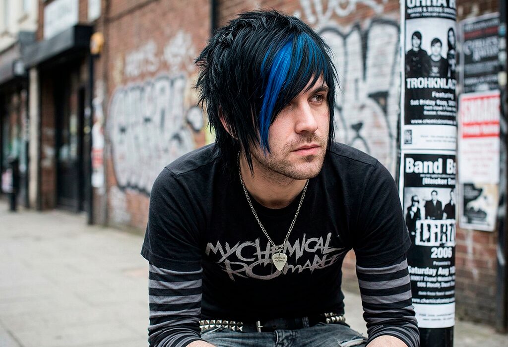Man with heavy emo fringe and blue streaks crouching beside graffiti-covered urban street.