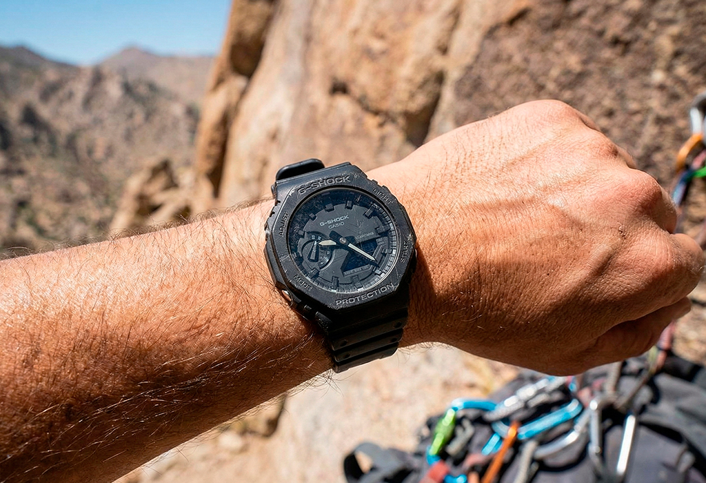 Black G-Shock on rugged rubber strap during rock climbing, carabiners blurred behind.