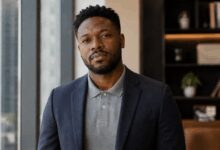 Black man wearing gray polo beneath navy suit jacket in modern office setting with confident expression.