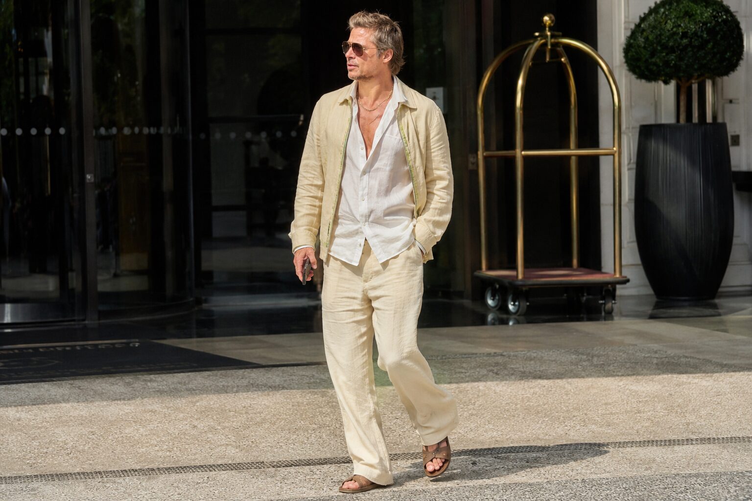 Brad Pitt in cream linen outfit wearing brown sandals outside a luxury hotel entrance.