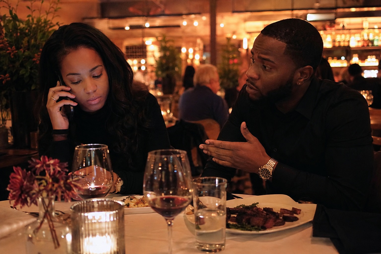 Black couple at a romantic restaurant dinner, with the woman distracted by a phone call while the man gestures and tries to speak to her.