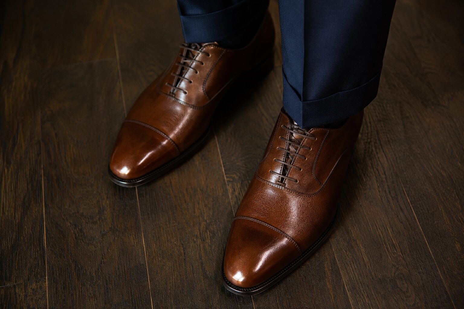 Polished brown leather dress shoes worn with navy trousers on dark wooden floor.