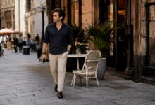 Man in navy shirt and cream trousers walks along café-lined street with phone.