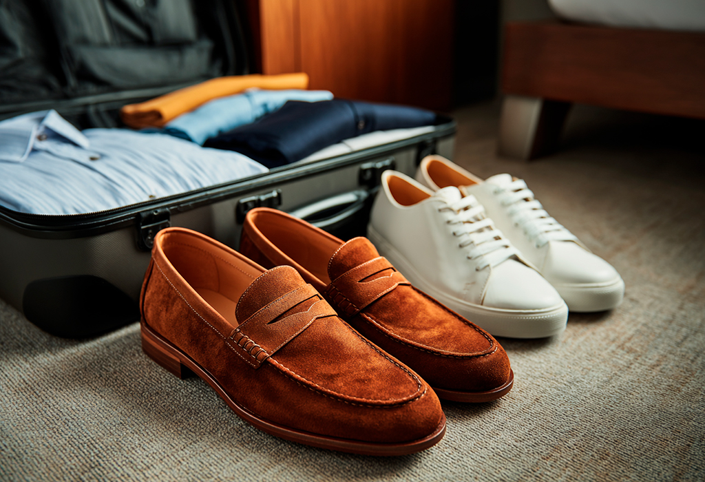 Brown suede loafers and white dress sneakers neatly placed beside an open suitcase with folded clothes