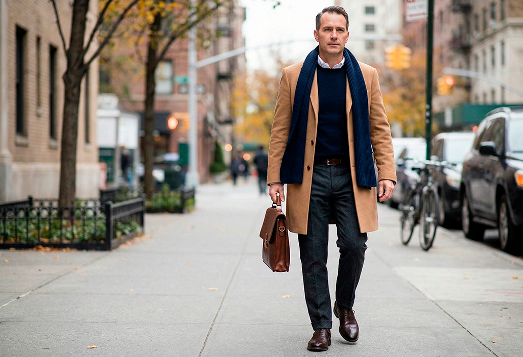 Smart casual winter outfit with a camel overcoat, navy sweater, grey trousers, and brown leather accessories