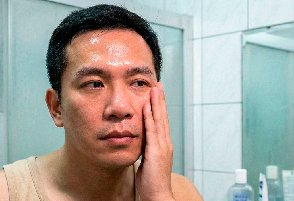 Man with oily, shiny skin touching cheek in bathroom mirror, checking texture and pores