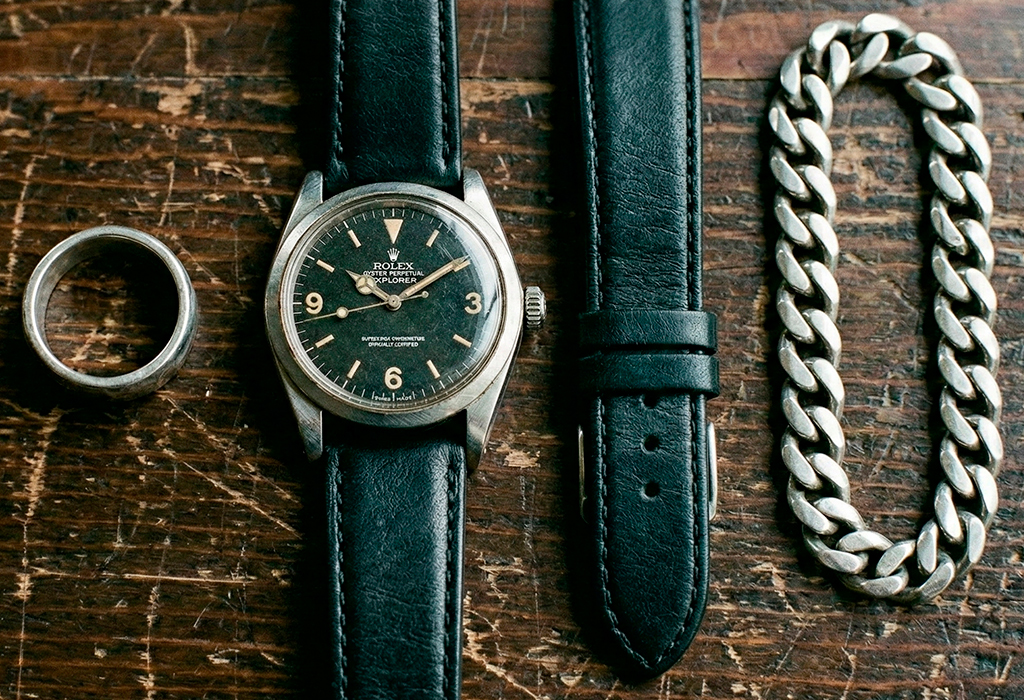 Flat lay of worn accessories on a wooden surface, including a watch with a leather strap, a metal ring, and a thick chain bracelet.
