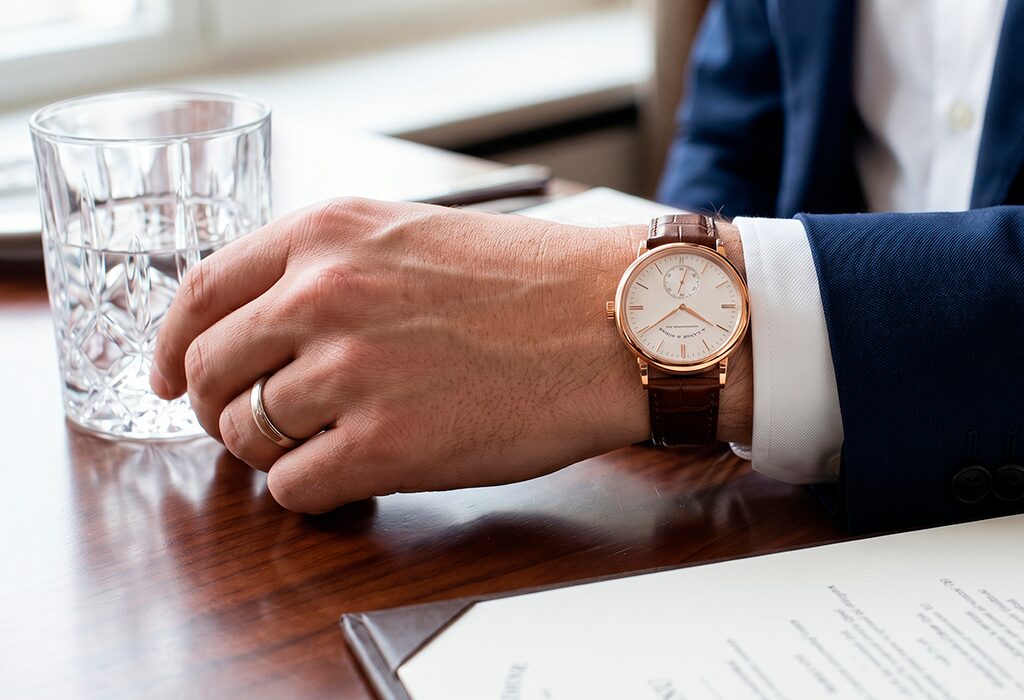 Rose gold dress watch with brown leather strap worn under navy suit sleeve.