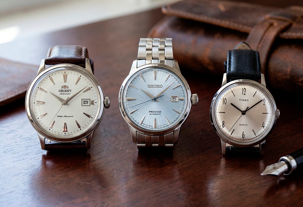 Three understated dress watches on leather and steel bracelets arranged on polished wood.