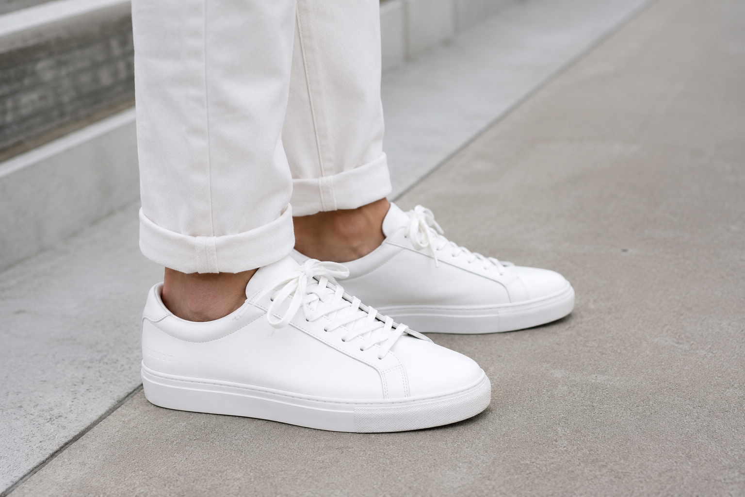 Close-up of a man wearing clean white Koio Capri sneakers with cuffed white trousers on a concrete walkway.