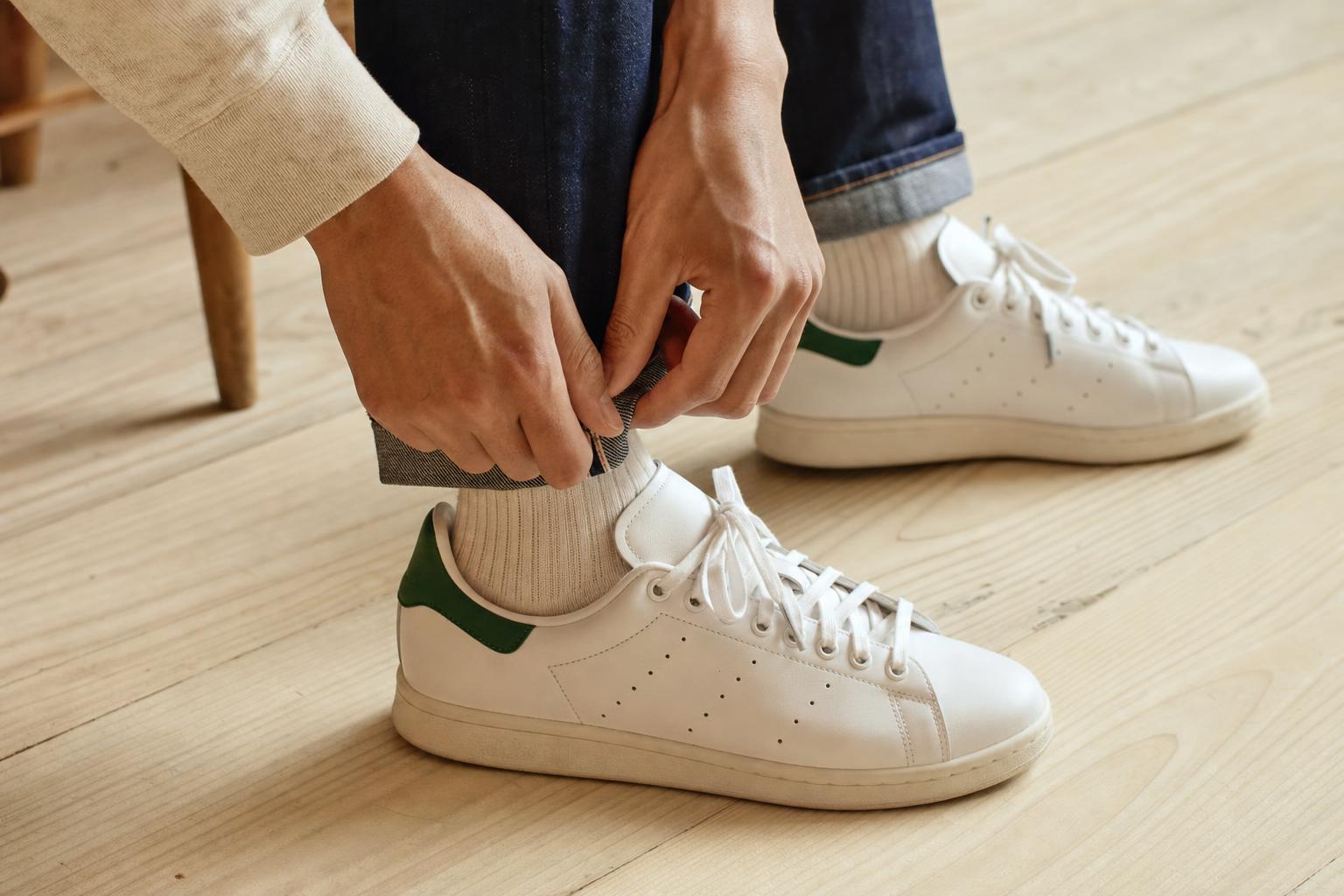 Close-up product shot of classic white Adidas Stan Smith leather sneakers with green heel tab, on a light wooden floor,